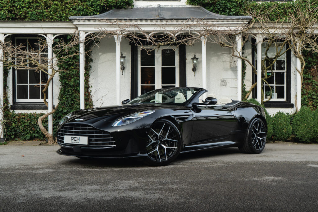 Aston Martin DB12 Volante hire in sleek black, parked in front of a luxury manor, showcasing its bold grille and elegant convertible design.